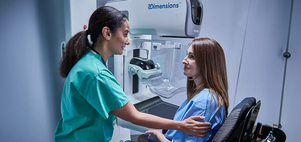 Femme assise devant un mammographe avec une professionnelle de santé à ses côtés.